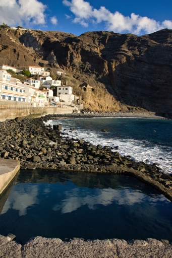 Una piscina tra gli scogli vicino a una fila di case. Sullo sfondo una scogliera altissima
