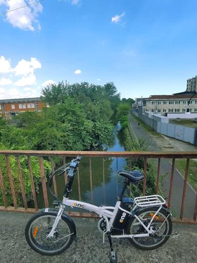 La bici assistita dell'autofficina: pieghevole bianca con le ruote da 20". La foto è presa da un ponte sotto cui si vede il naviglio Martesana, della vegetazione, un edificio commerciale sulla destra. Il cielo è azzurro con qualche nuvola