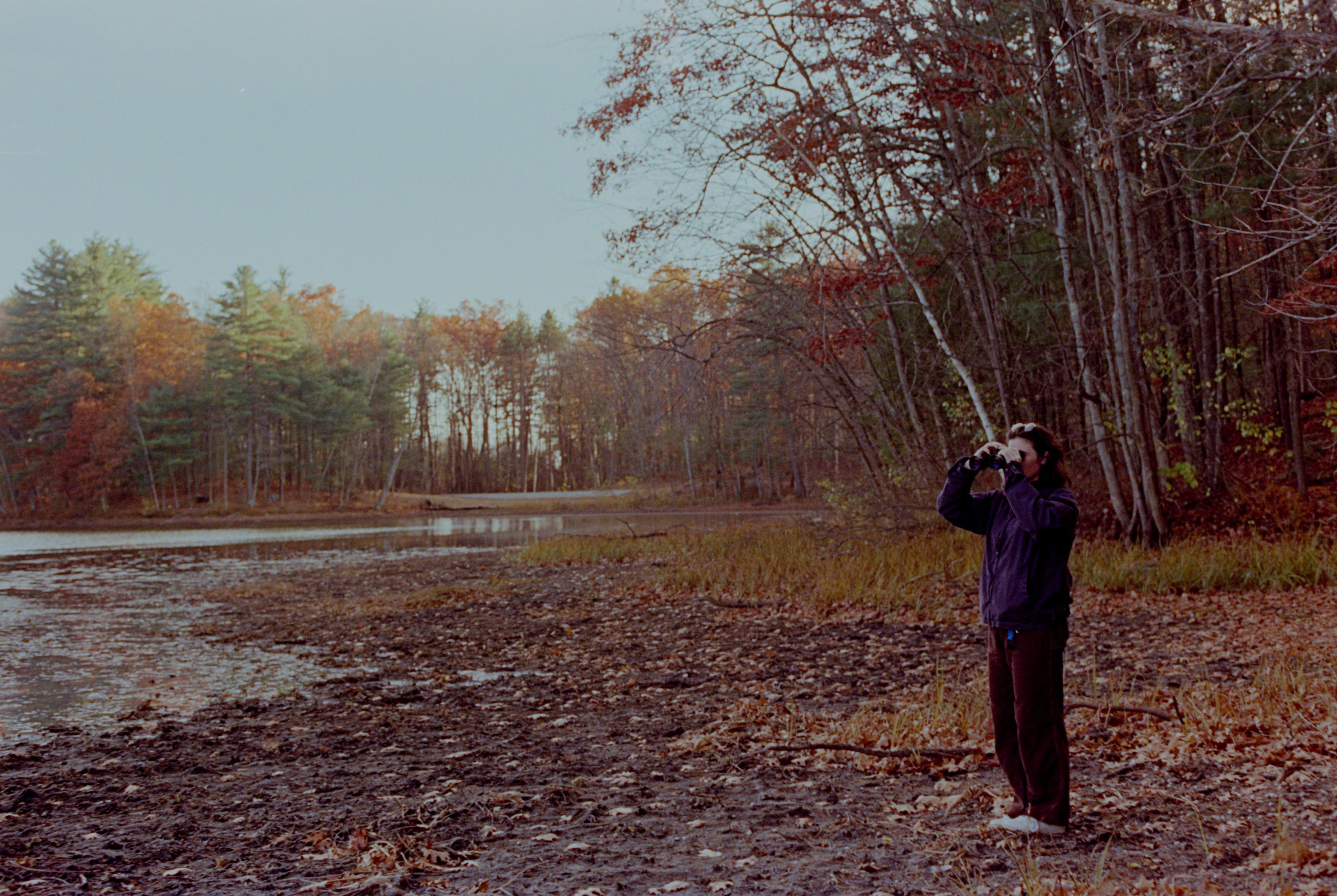 A person stands on the dried shore of a pond, peers through a set of binoculars. It is fall. 