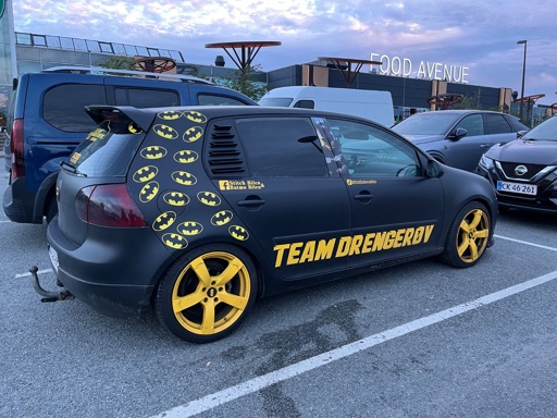 Bilen på billedet er en sort personbil med iøjnefaldende gule fælge og flere Batman-logoer på siden. Den bærer teksten "TEAM DRENGERØV" i store gule bogstaver, hvilket giver den et unikt og tematisk udtryk. Bilen er tydeligt dekoreret med et superhelte-inspireret design og skiller sig markant ud i bybilledet.