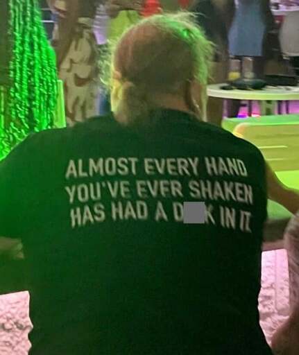 A person is wearing a black t-shirt that has white lettering on the back. The back of the t-shirt reads "Almost every hand you've ever shaken has had dick in it." The word dick is rudely censored.