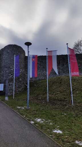 Four flags hoisted vertically, each on their own flagpoles. From left to right, those flags are of: European Union, Slovakia, Bratislava and Bratislava City Museum. In front of the image, a part of the asphalt can be visible. Behind it is a grass surface followed by a gray stone wall behind the flags.
