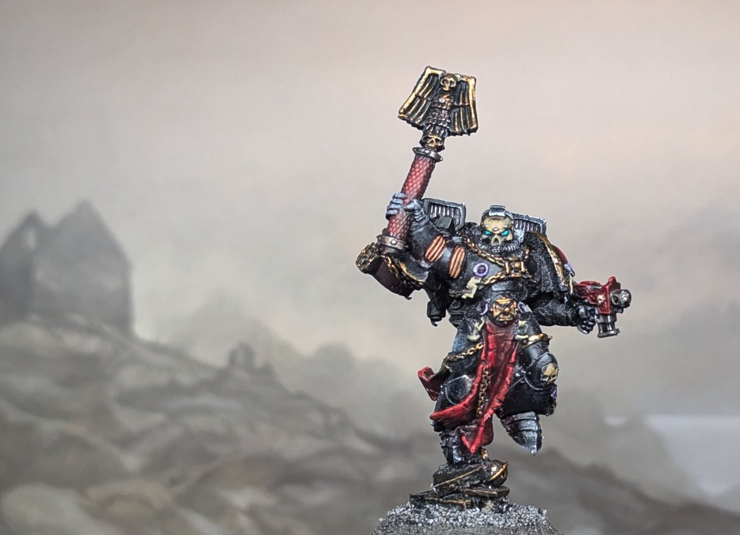 A painted Warhammer 40k miniature, a Chaplain with Jump Pack, featuring black armor with red cloth and lots of gold details. In front of a painted backdrop of a misty hillside with ruined cottage.