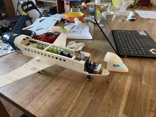 Su un tavolo di legno chiaro, c'è un aereo di Lego bianco e blu in fase di costruzione.  Una parte della fusoliera è aperta, mostrando i sedili interni. Vicino all'aereo, c'è una parte del muso del velivolo separata e un computer portatile.  Sullo sfondo si intravedono altri oggetti, ma non sono dettagliati.